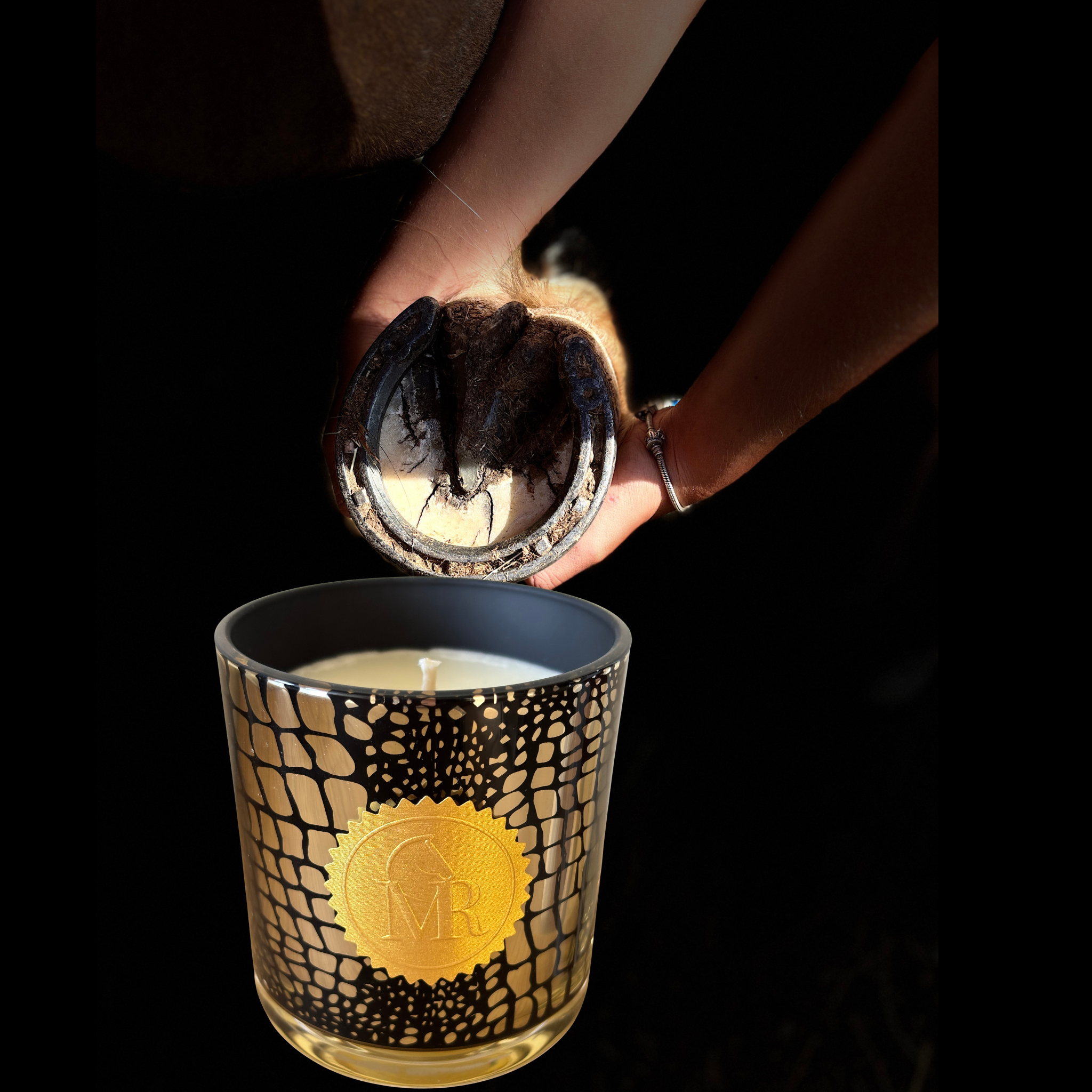 Miss Rio English Pear & Freesia luxury scented candle in a black and gold patterned glass jar with a gold Miss Rio seal, photographed with hands holding a horseshoe above the candle.