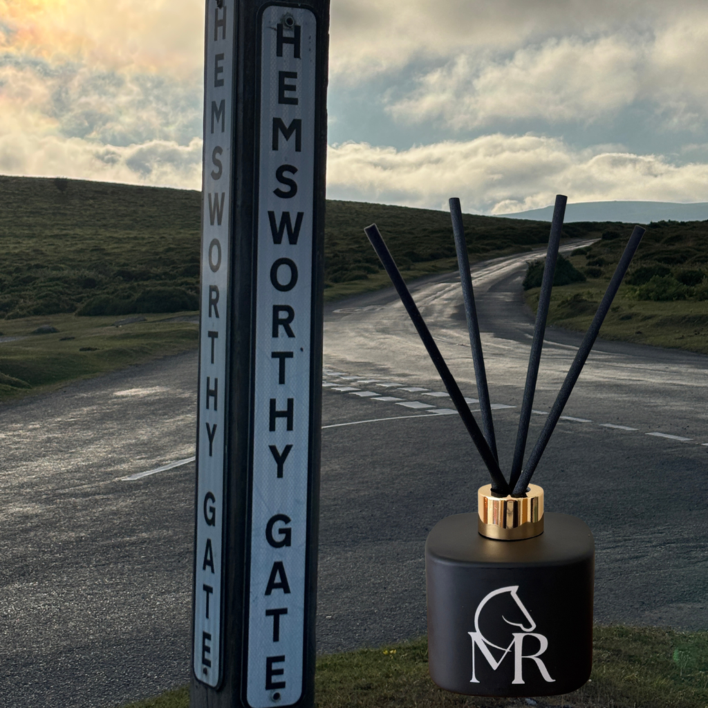 Miss Rio Dartmoor Smokey Embers luxury reed diffuser in a matte black vessel with gold cap and MR equestrian horse logo, placed beside the Hemsworthy Gate road sign on Dartmoor with a dramatic moorland road stretching into the distance. Born on Dartmoor. missrio.co.uk