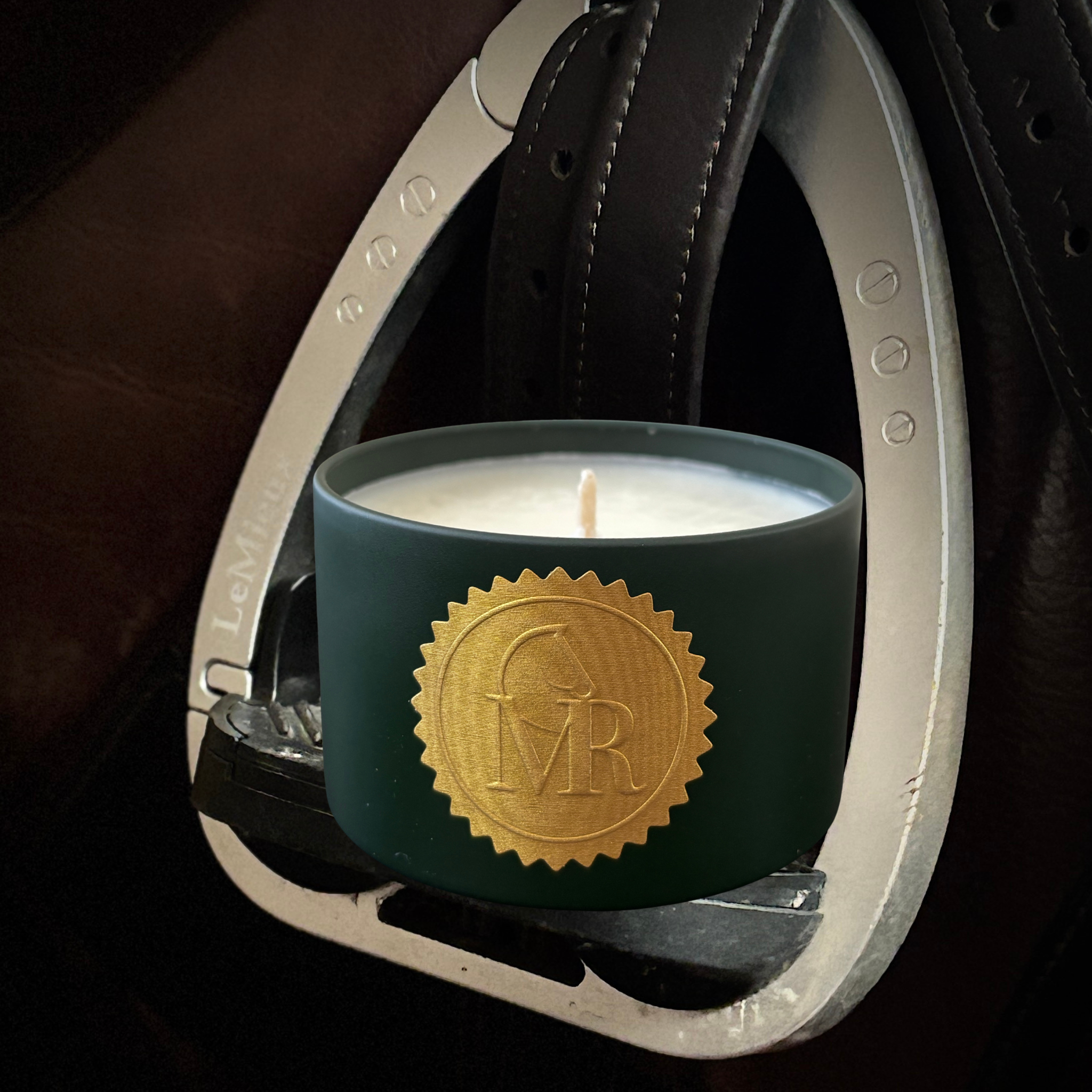 Miss Rio Tomato Vine candle in a dark green tin with a gold horse seal, displayed inside a white riding stirrup.”