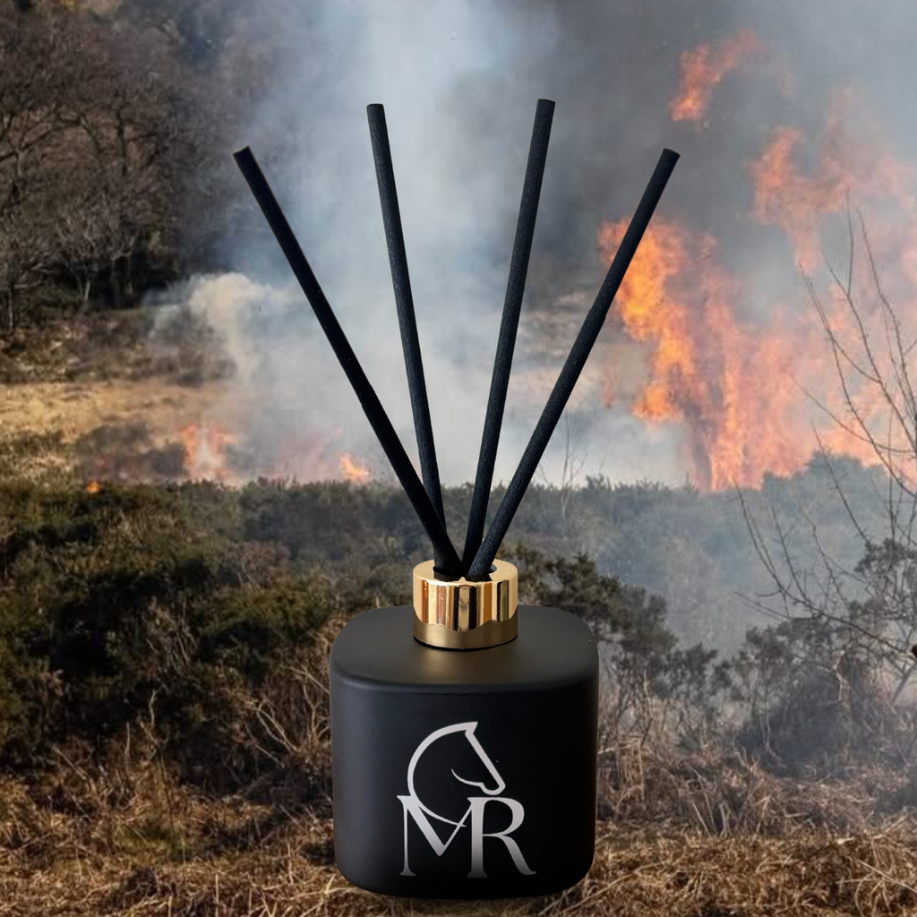 Miss Rio Dartmoor Smokey Embers luxury reed diffuser in matte black with gold collar and black reeds, set against a dramatic moorland controlled burn with flames and smoke on Dartmoor