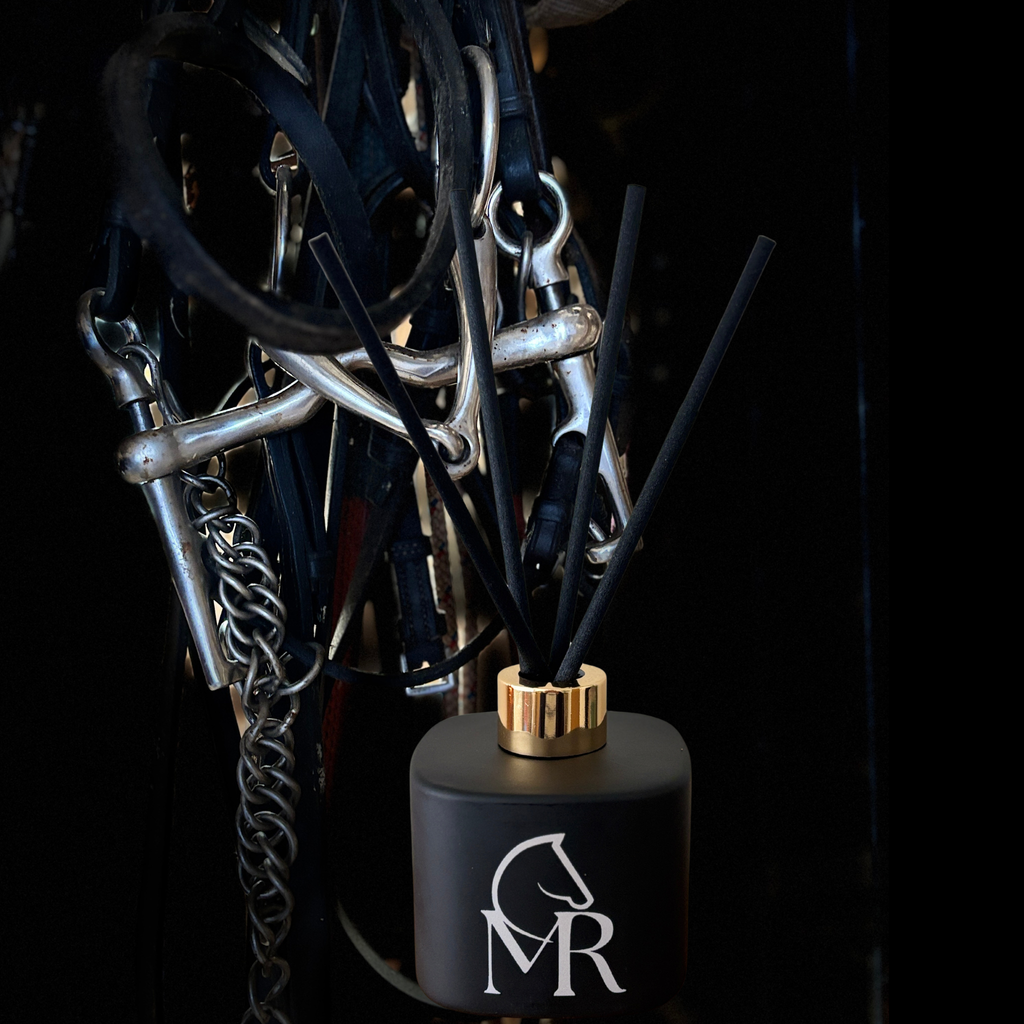 Miss Rio luxury reed diffuser in matte black with gold collar and black reeds, styled against equestrian bridles, bits and leather tack in a dark tack room setting